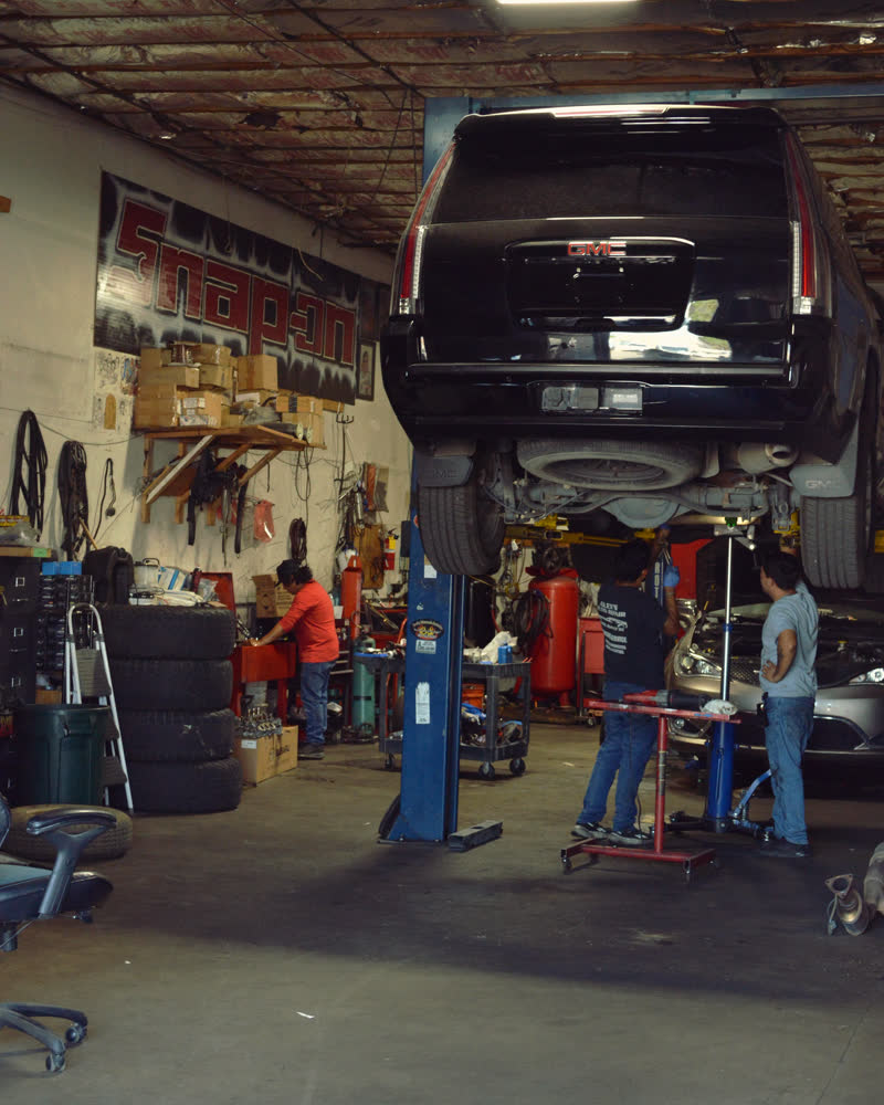 The shop floor: an SUV up on the two-post lift and three people working at benches and cars around it.